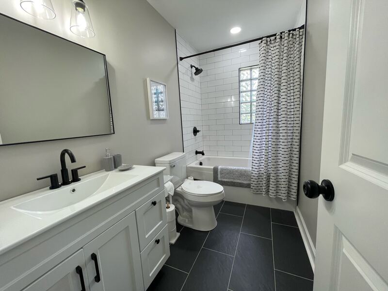A bathroom with a white vanity, black fixtures, and a bath and shower lined in white subway tile with a window above.