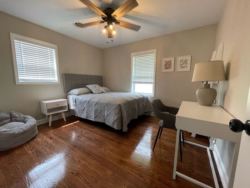 A bedroom with a grey upholstered headboard, a white nightstand and desk, and a round pet bed in the corner.