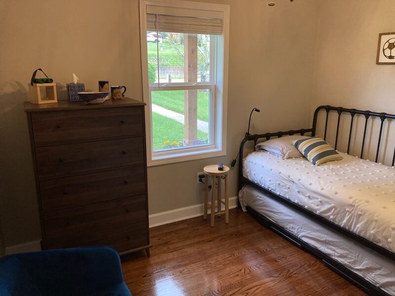 A bedroom with a black metal daybed and its trundle bed pulled out underneath, a tall dark-wood dresser, and a window looking onto the front lawn.