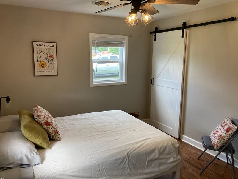 A bedroom with white bedding, olive and floral cushions, a framed Picasso print beside the window, and a sliding barn door to the closet.