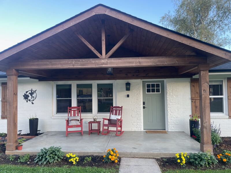 The covered front porch, framed by heavy stained-timber posts and a gabled roof, with two red rocking chairs and a small table between them, and the sage-green front door beyond.